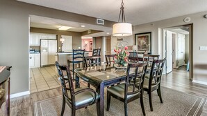 A spacious dining area with a glass-top table surrounded by wooden chairs, a hanging light fixture, and a view into the kitchen area. - Enjoy Tranquility at Barefoot Resort & Yacht Club 7th Floor Waterfront Unit (North Myrtle Beach)