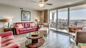 A spacious living room with a red leather sofa, a white armchair, a glass coffee table, and a ceiling fan. Large windows offer a view of the cityscape. - Enjoy Tranquility at Barefoot Resort & Yacht Club 7th Floor Waterfront Unit (North Myrtle Beach)