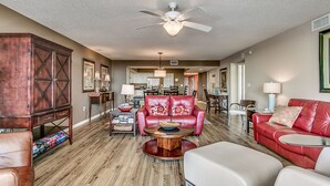 A spacious living room with a red leather sofa, a white armchair, and a wooden coffee table. A ceiling fan is visible, and the room has hardwood flooring. - Enjoy Tranquility at Barefoot Resort & Yacht Club 7th Floor Waterfront Unit (North Myrtle Beach)