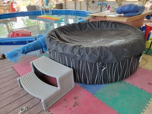 A hot tub with a black cover, a white chair, and a colorful mat on a wooden deck.
