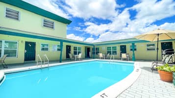 Uma área de piscina com piscina azul cristalina, área de estar coberta e um edifício com portas numeradas.