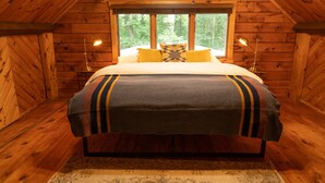Een houten slaapkamer met een bed, twee bedlampjes en een raam met uitzicht op een bos.