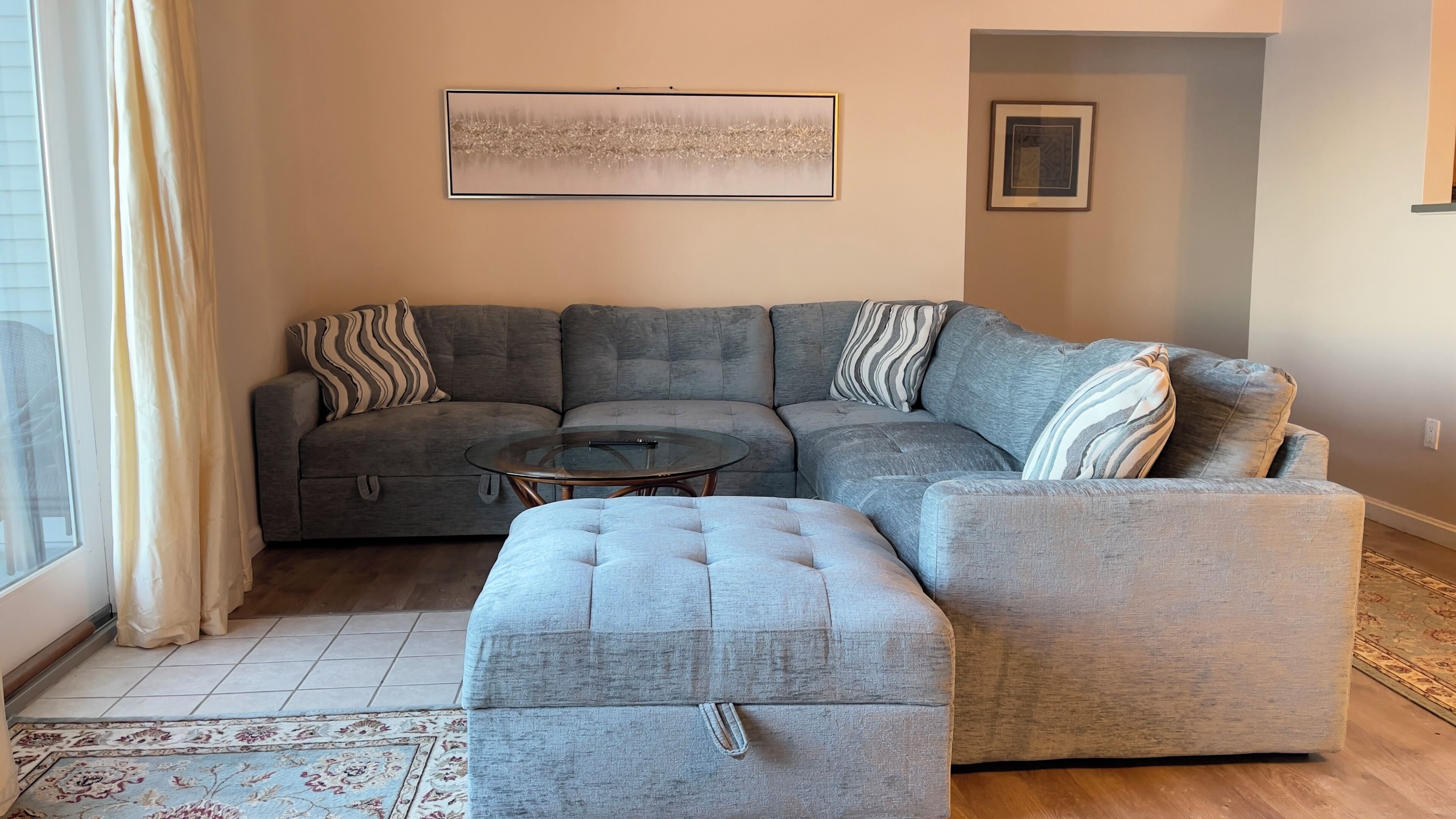 Un salon avec un canapé d’angle gris, une table basse en verre et un tapis à motifs.