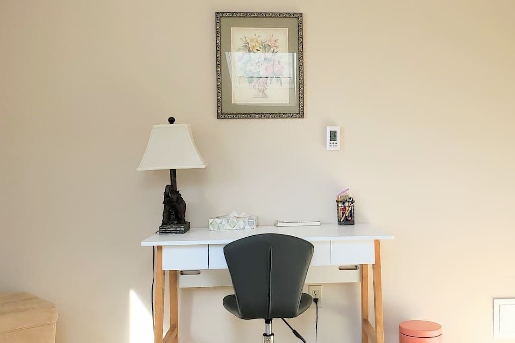 Een wit bureau met een zwarte stoel, een lamp, een ingelijst beeld en een kleine, roze prullenbak.