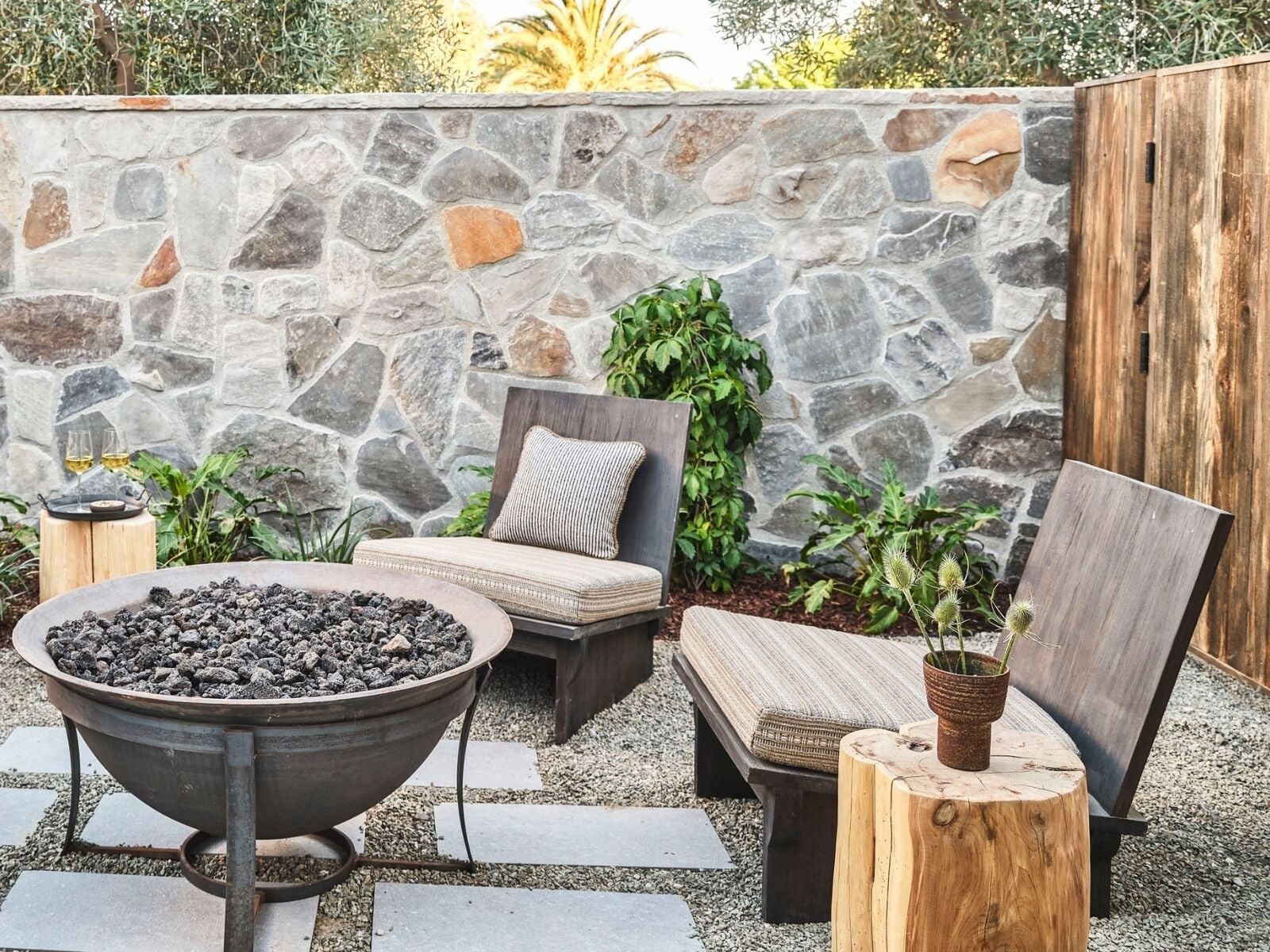A stone wall, outdoor seating with cushions, a metal fire pit, and potted plants.