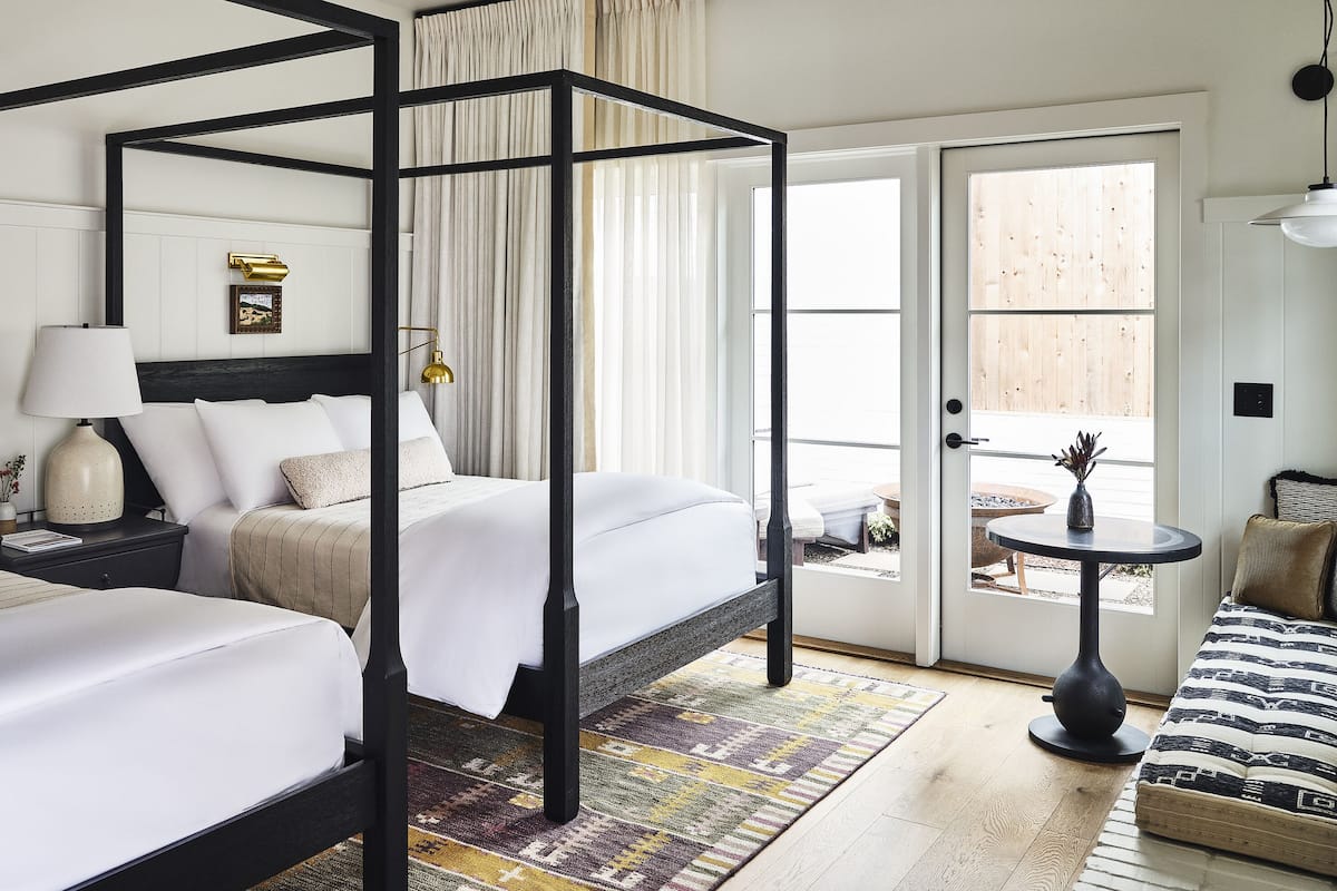 A four-poster bed with white linens, a bedside table with a lamp, and a view of the outdoor patio.