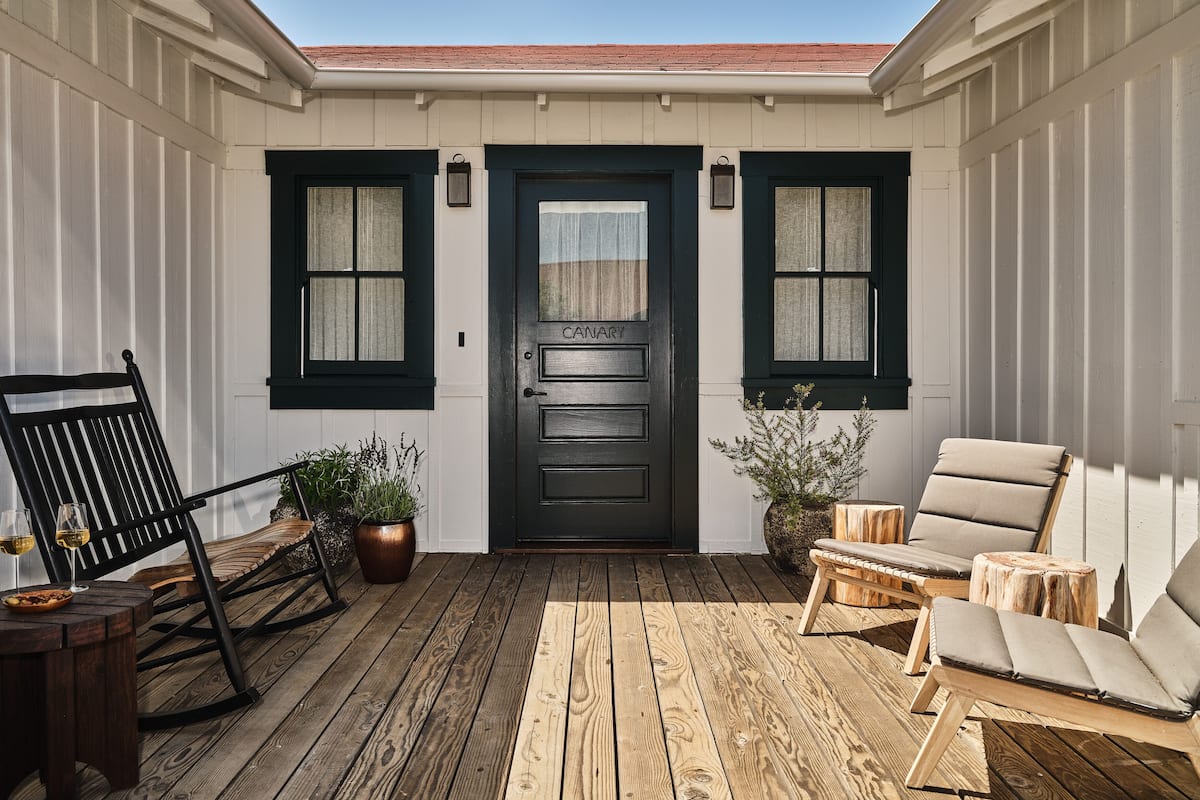 A wooden deck with a black door labeled "CANARY," two potted plants, a wooden rocking chair, and two lounge chairs.