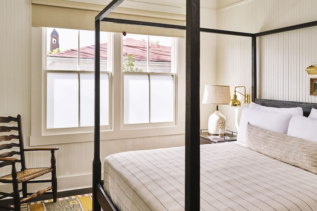 A four-poster bed with a white and black frame, a wooden chair, a nightstand with a lamp, and a window with a view of a building and a red roof.