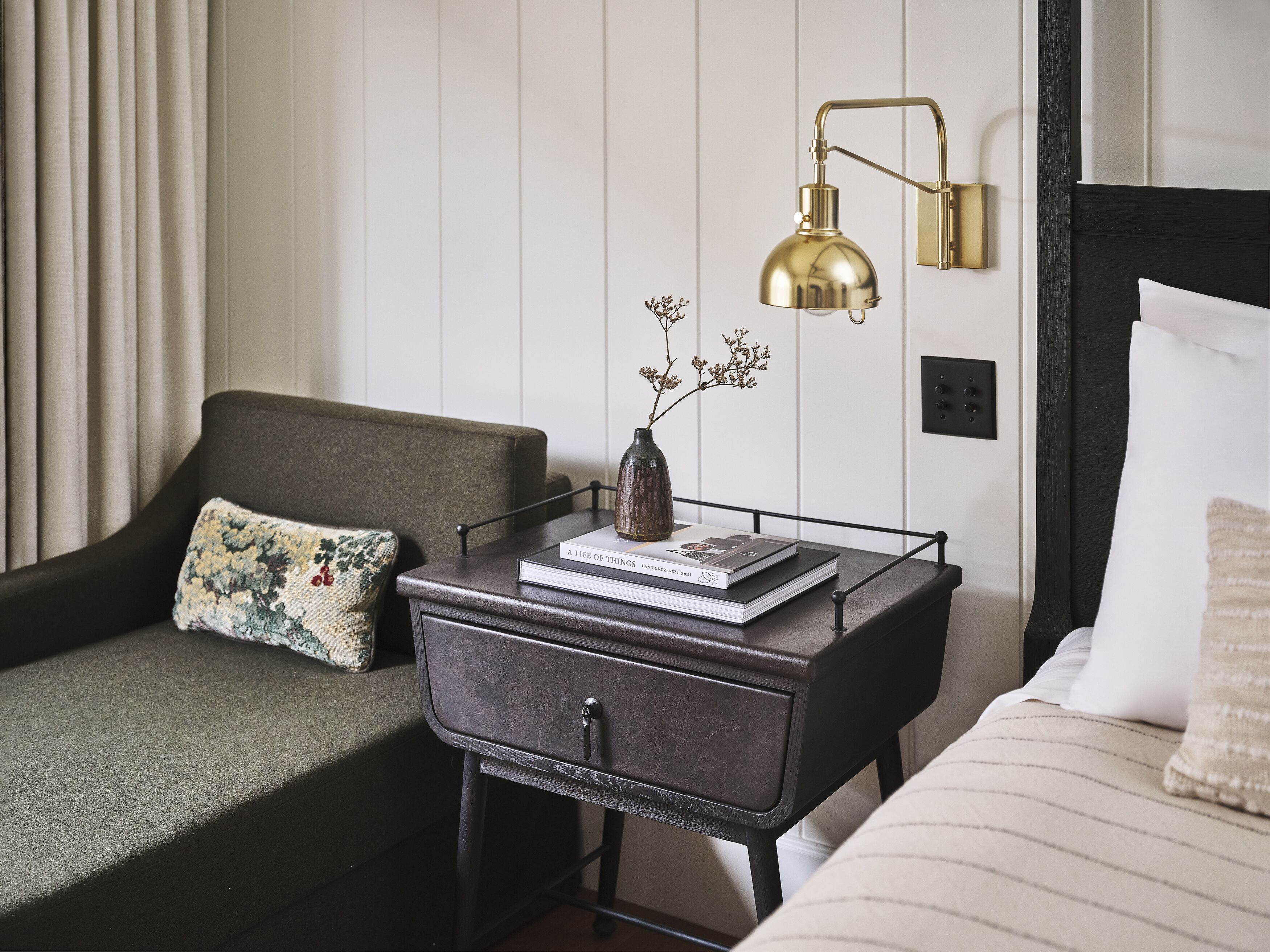 A neatly arranged hotel room with a bed, a bedside table with a lamp, a sofa, and a small side table with a book and a vase.