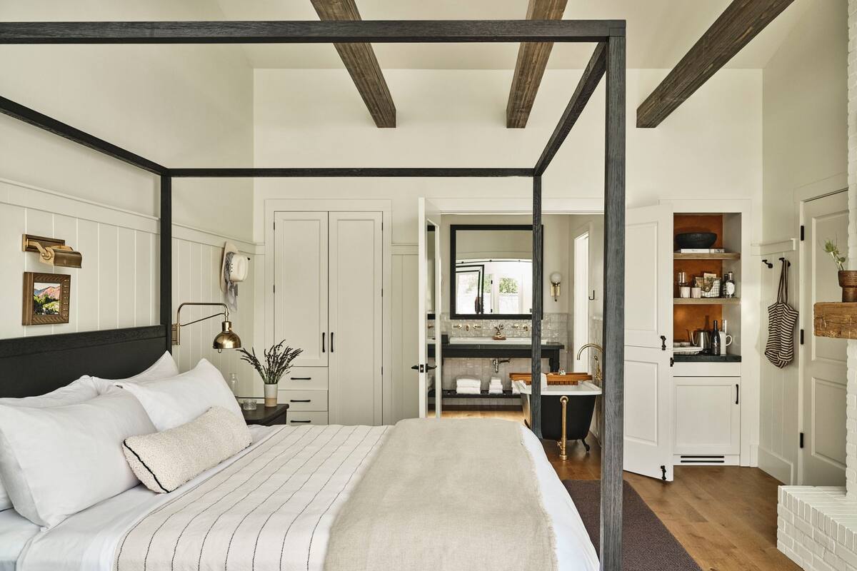 A four-poster bed with a headboard, a bedside table with a lamp, and a nightstand. There is a bathroom with a sink, mirror, and towel rack. The room has wooden beams on the ceiling and white walls.