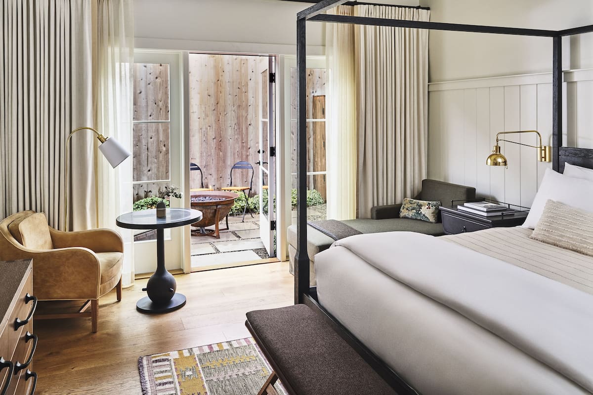 A modern hotel room with a large bed, a desk, a chair, and a small table. There is a balcony with a view of the outdoors.