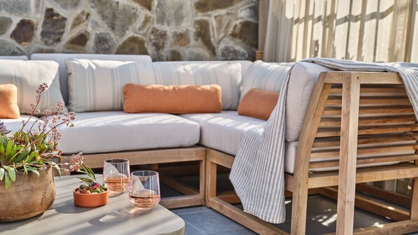 A cozy outdoor seating area with a cushioned bench, a wooden table with glasses, and potted plants.