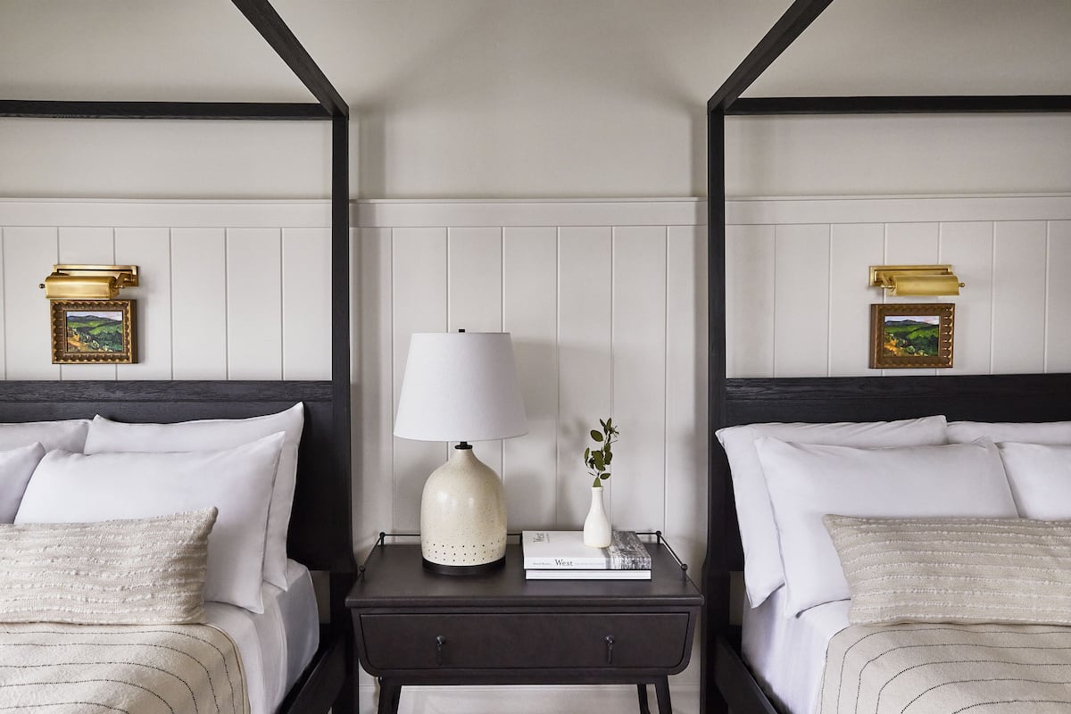 A hotel room with two beds, a nightstand with a lamp, a book, and a vase with a plant.