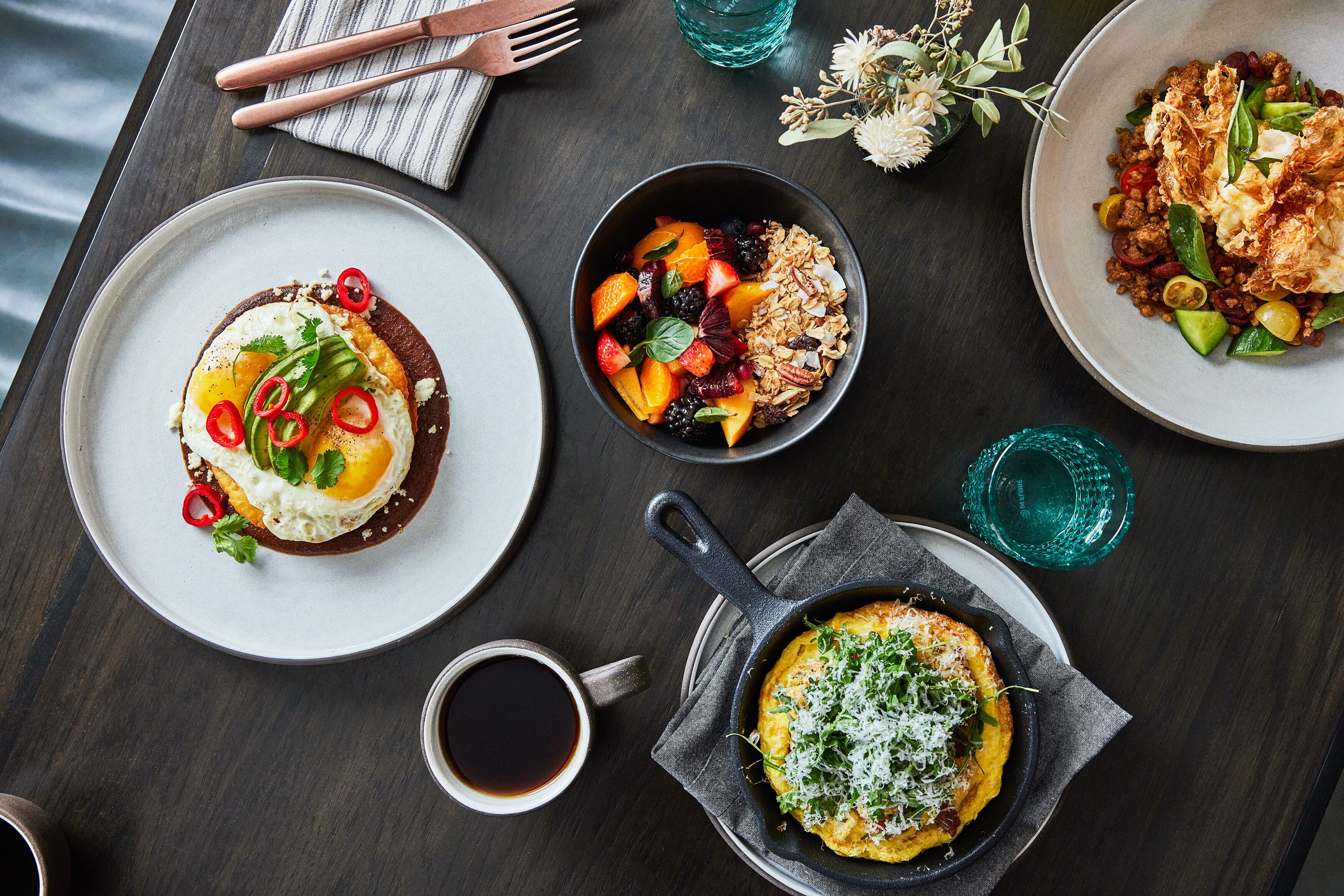 A dining table with various dishes, including a plate with a fried egg, a bowl of fruit salad, and a skillet with a baked dish.