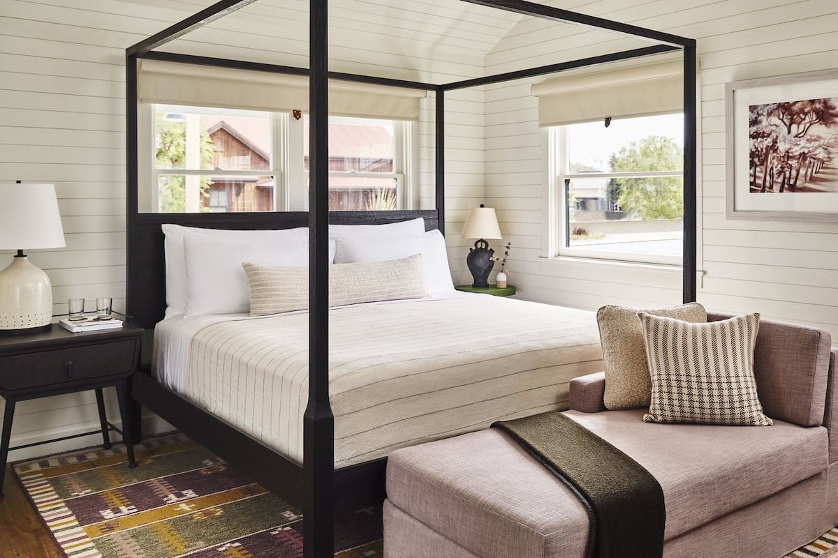 A four-poster bed with white linens, a nightstand with a lamp, a chair with patterned cushions, and a framed picture on the wall.