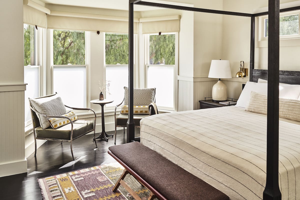 A four-poster bed with a striped bedspread, two upholstered chairs with patterned cushions, a small round table with a vase, and a nightstand with a lamp and a framed picture.