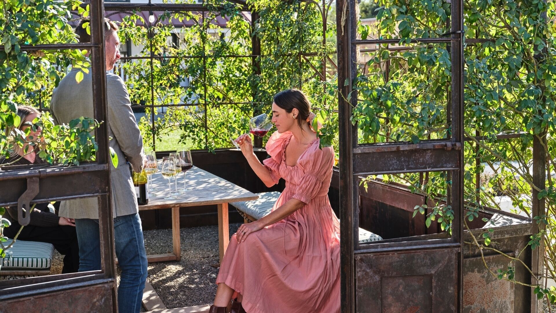 A woman in a pink dress sitting at a table with wine glasses, surrounded by greenery and a metal structure.