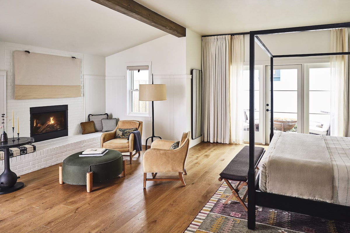 A spacious bedroom with a four-poster bed, a fireplace, a seating area with a chair and ottoman, and a view of the outdoors through large windows.