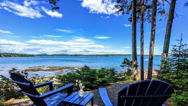 Dos sillas Adirondack y una mesita con un florero, ubicadas sobre un área de grava con vista a un lago, árboles y una orilla lejana.