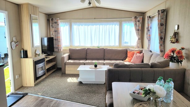 A cozy living room with a sofa, coffee table, and a TV. There is a fireplace, a window with curtains, and a ceiling fan. - St Osyth- Sleeps8-freeparking-wifi-holidaypark (Clacton-on-Sea)
