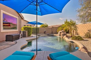 Une piscine dont les bords sont bordés de pierres, un espace détente avec des coussins bleus et un grand parasol bleu offrant de l’ombre.