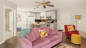 Een moderne woonkamer met een roze bank, een rode fauteuil en een geel poufje. In de achtergrond bevindt zich een keuken met witte kasten en een plafondventilator.