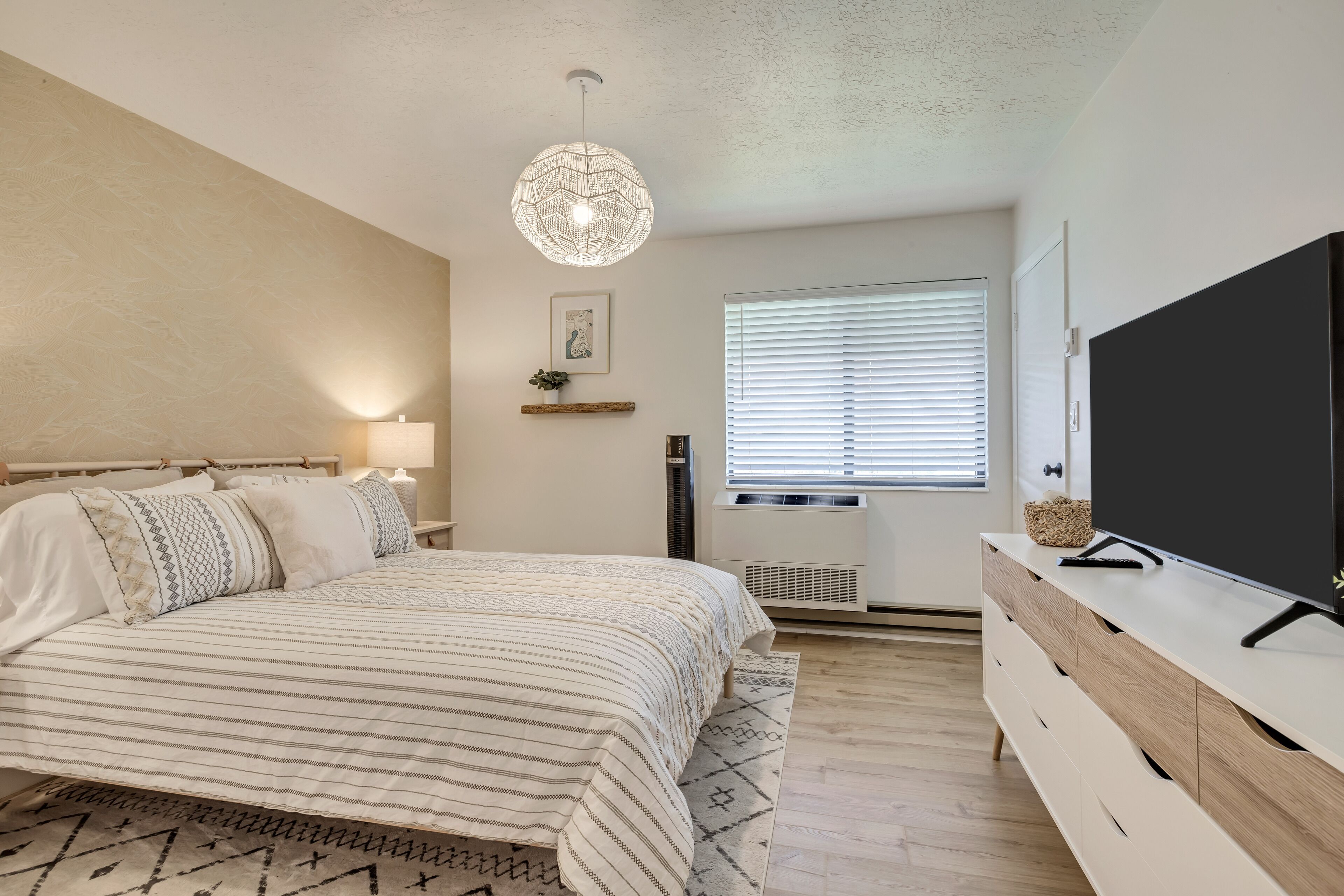 A bedroom with a bed, bedside table, lamp, TV, and a window with blinds.