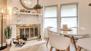 A modern living room with a fireplace, white walls, and a round mirror. There is a dining table with chairs and a potted plant.