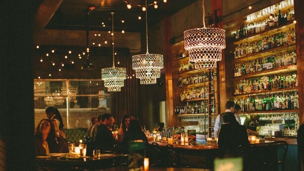 A bar with hanging pendant lights, shelves of liquor bottles, and patrons seated at tables.