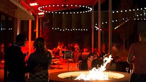 A restaurant with a central fire feature, outdoor seating, and string lights.