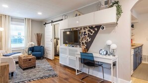A modern living room with a sofa, a desk, and a TV. There is a window with curtains, a rug, and a plant.