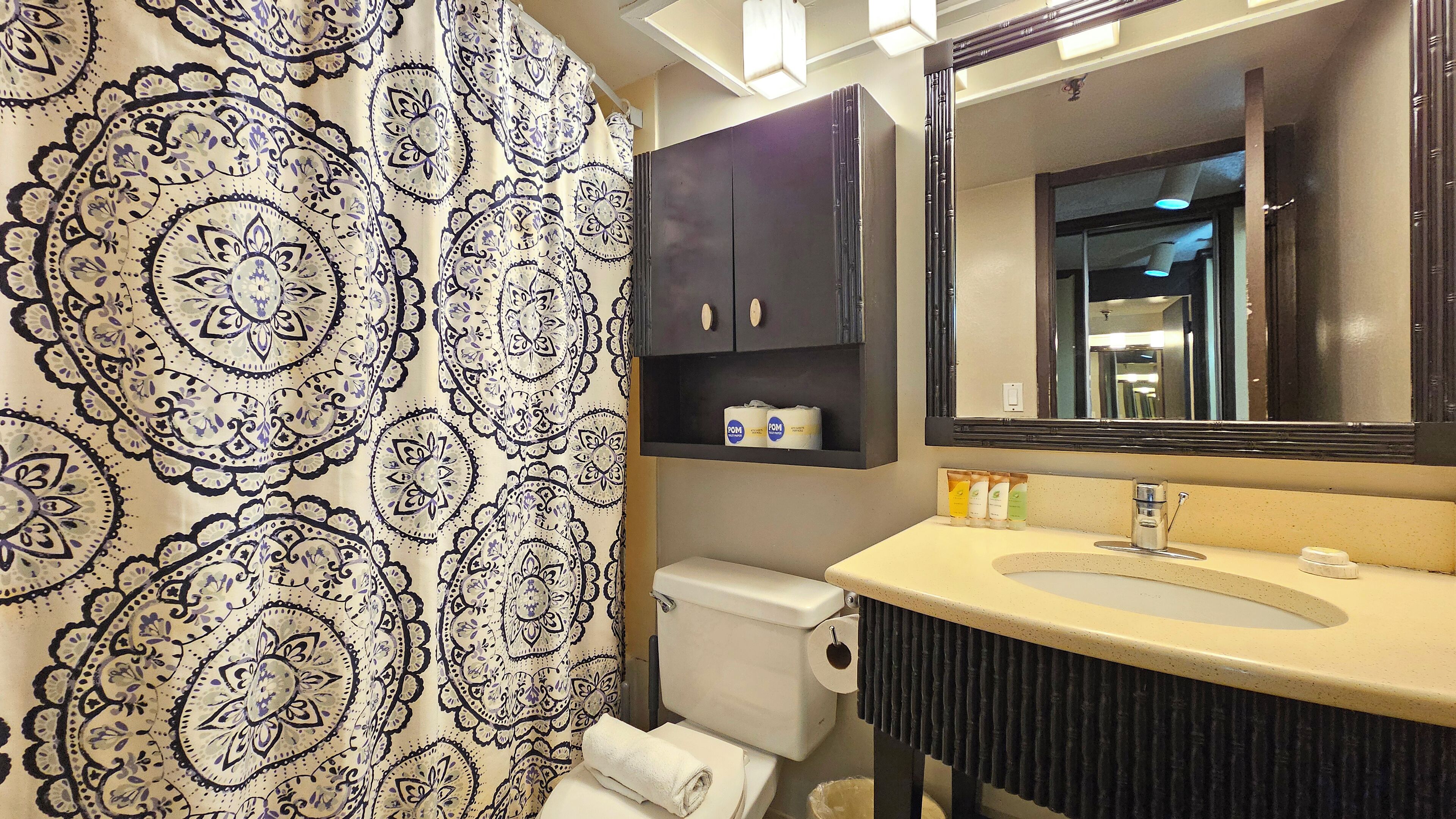 Un baño con cortina de ducha estampada, un inodoro, un lavabo con artículos de aseo y un espejo sobre el lavabo.