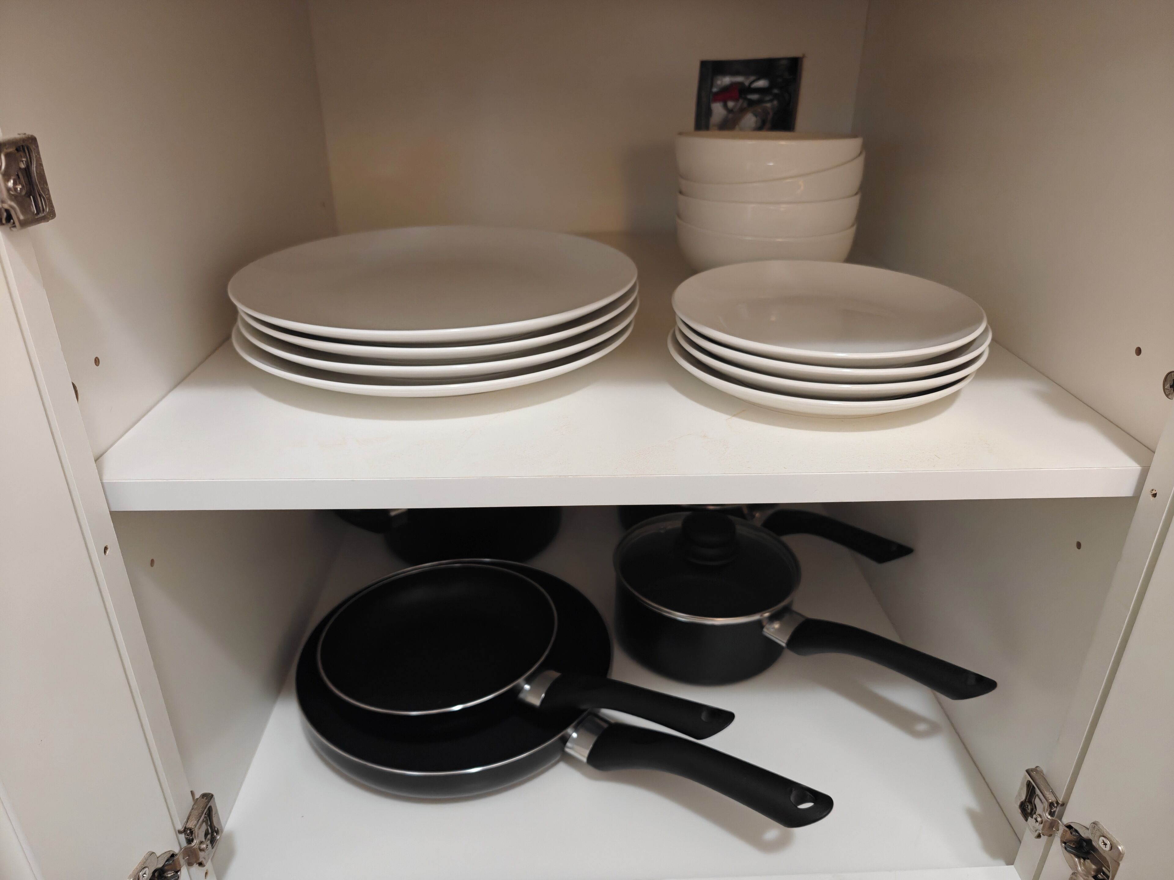 A kitchen cabinet with white shelves holding stacked plates and a black frying pan with a pot underneath.
