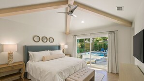 Un dormitorio con una cama grande, ventilador de techo, mesita de noche, banco, televisor y vistas a un área de piscina exterior.