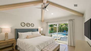 A bedroom with a large bed, a ceiling fan, a nightstand, a bench, a TV, and a view of an outdoor pool area.