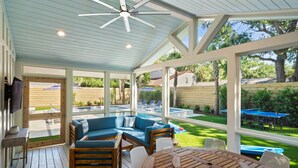 Un área de estar exterior cubierta con piscina, mobiliario de madera y ventanas amplias.