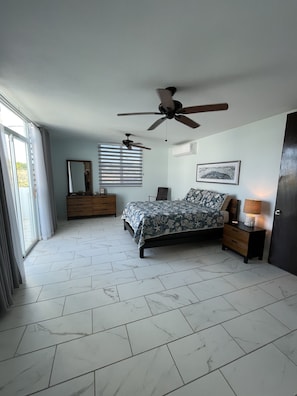 Une chambre à coucher comprenant un lit, une commode, une table de chevet, un ventilateur de plafond, une fenêtre avec des stores et une porte.