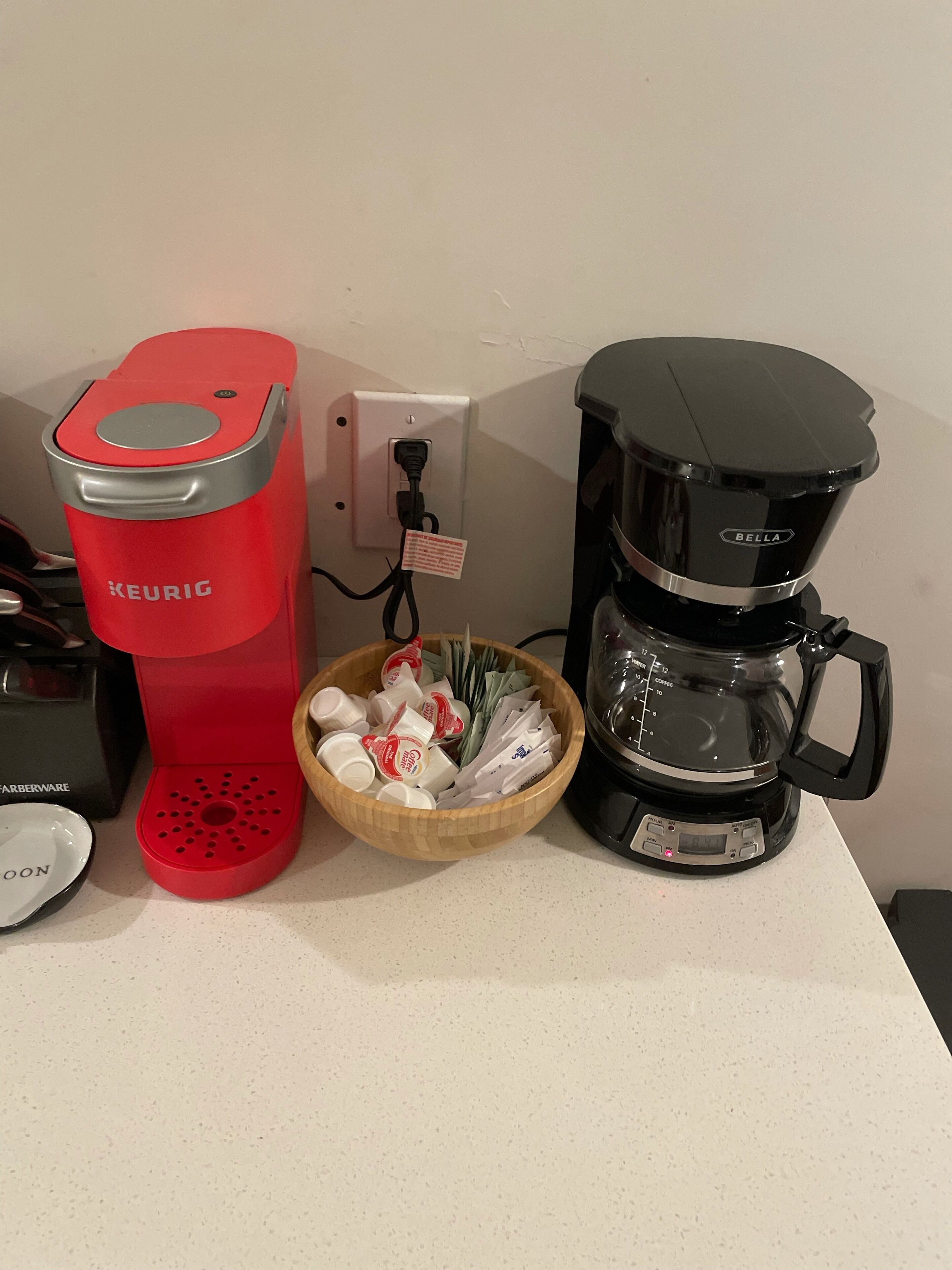 A red Keurig coffee maker, a black coffee maker, and a bowl of coffee pods on a countertop.