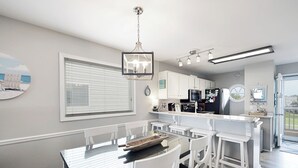 A modern kitchen with white cabinetry, a central island, and a dining area with a glass table and white chairs. - "Waters Edge 429-D" - Oceanfront Condo w/ Community Pool, 2 Bedrooms / 2 Bathrooms (Holden Beach)