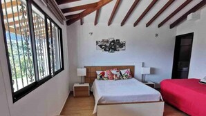 A bedroom with a large bed, a smaller bed, a nightstand, a red bedspread, and a window with metal bars. - Country house surrounded by nature  (El Carmen de Viboral)