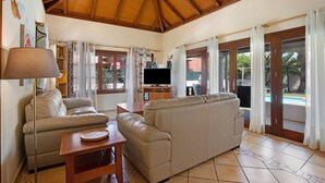 A living room with a wooden ceiling, a sofa, a television on a stand, and a view of a swimming pool through glass doors.