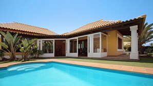 A house with a swimming pool, a covered patio, and a garden with palm trees.