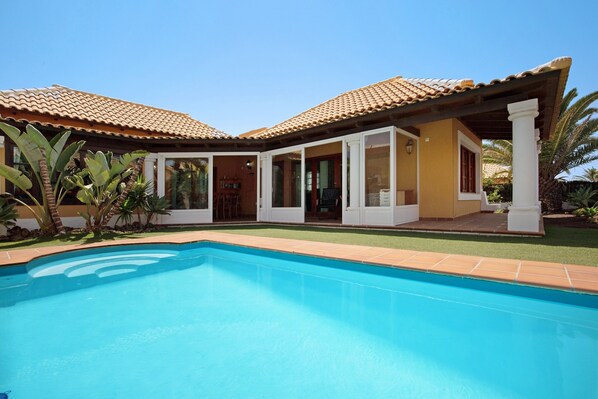 A house with a swimming pool, a covered patio, and a garden with palm trees.