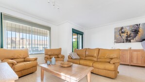 Un salon spacieux avec une grande fenêtre, un ensemble de canapés beige, une table basse en bois et un tableau accroché au mur.
