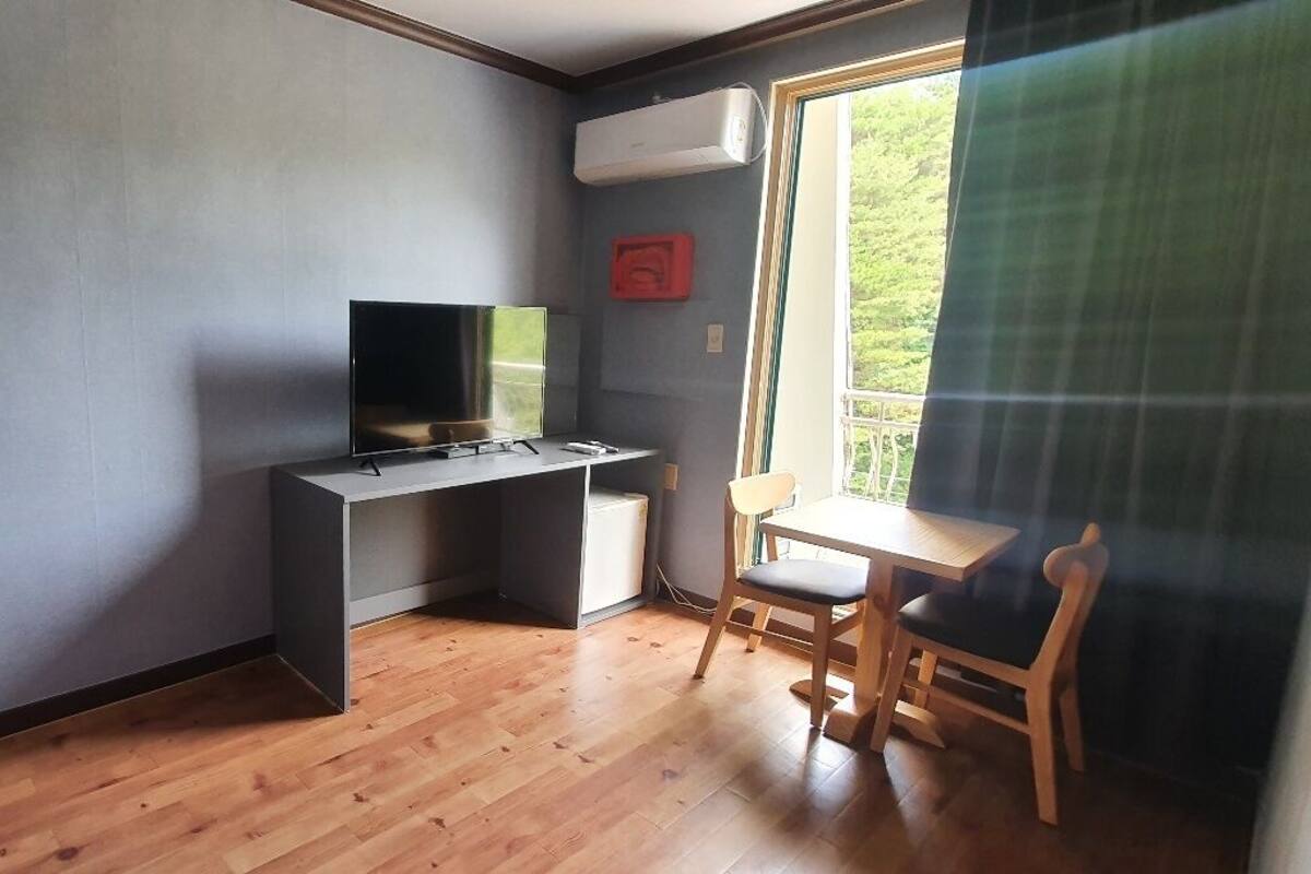Una habitación con un escritorio de madera, una silla, un televisor y una ventana con vistas a zonas verdes.