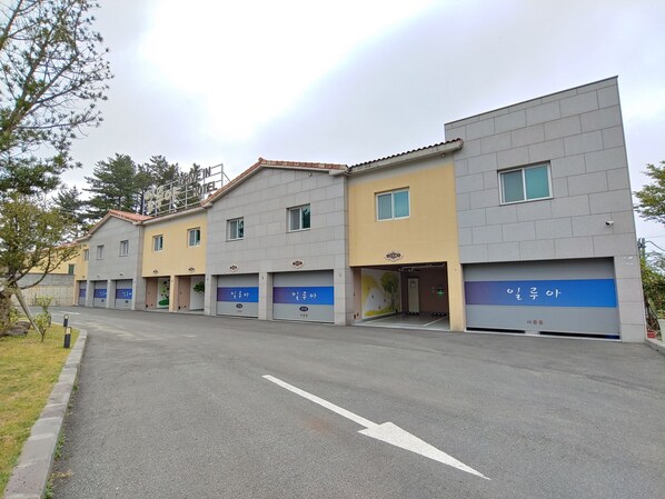 A row of hotel buildings with multiple parking spaces and a clear road in front. - Jeju Ilua Hotel (Jeju City)