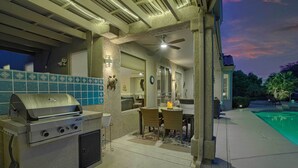 An outdoor kitchen with a grill, dining area with chairs, and a pool in the background. - Pool & Golf Cart: Sun City Grand Home! (Surprise)