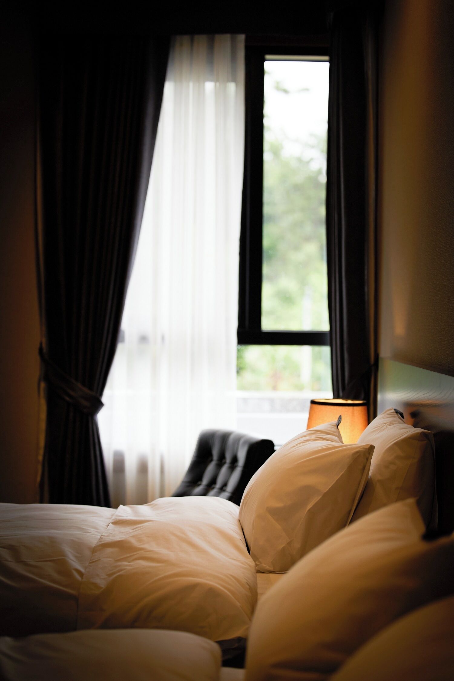 Una cama con ropa blanca, una lámpara en el lateral y una ventana con cortinas.