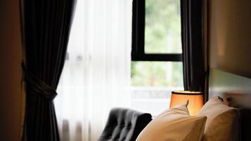 Una cama con ropa blanca, una lámpara en el lateral y una ventana con cortinas.