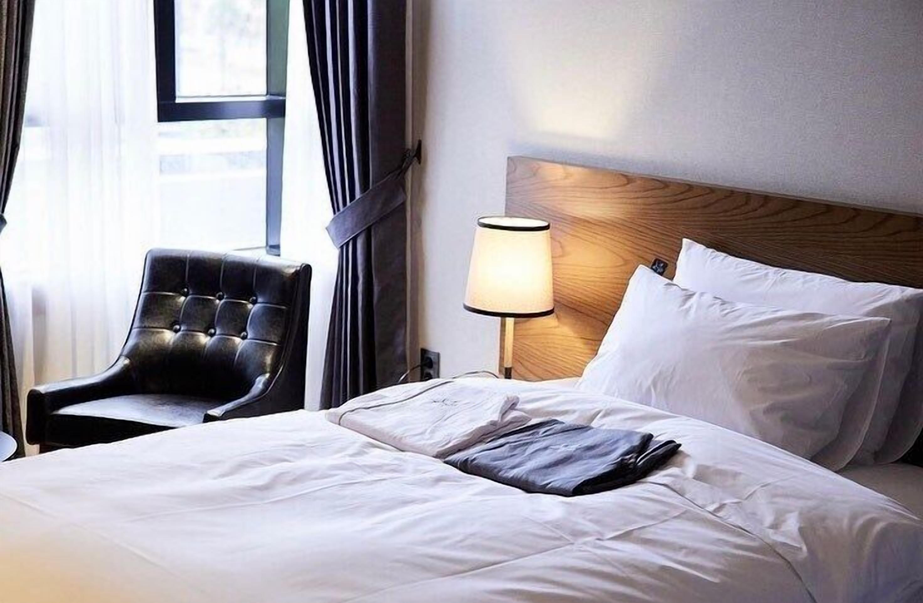 A hotel room with a bed, a chair, a lamp, and a window with curtains.